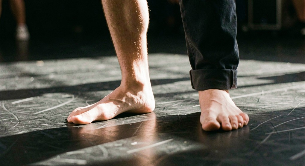 Pied nu appuyé fortement sur un plancher de danse noir, orteils écartés, jambe fléchie, lumière rasante venant du sol, cadrage vertical très serré