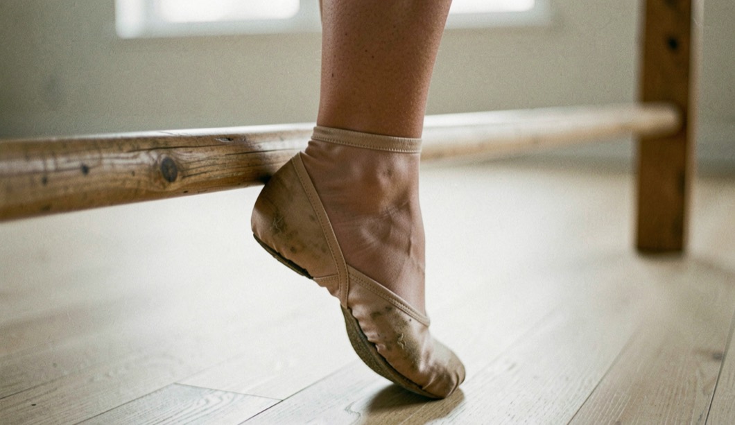 Cou-de-pied tendu en demi-pointe près d’une barre en bois, chausson de satin usé, parquet clair, lumière douce de studio