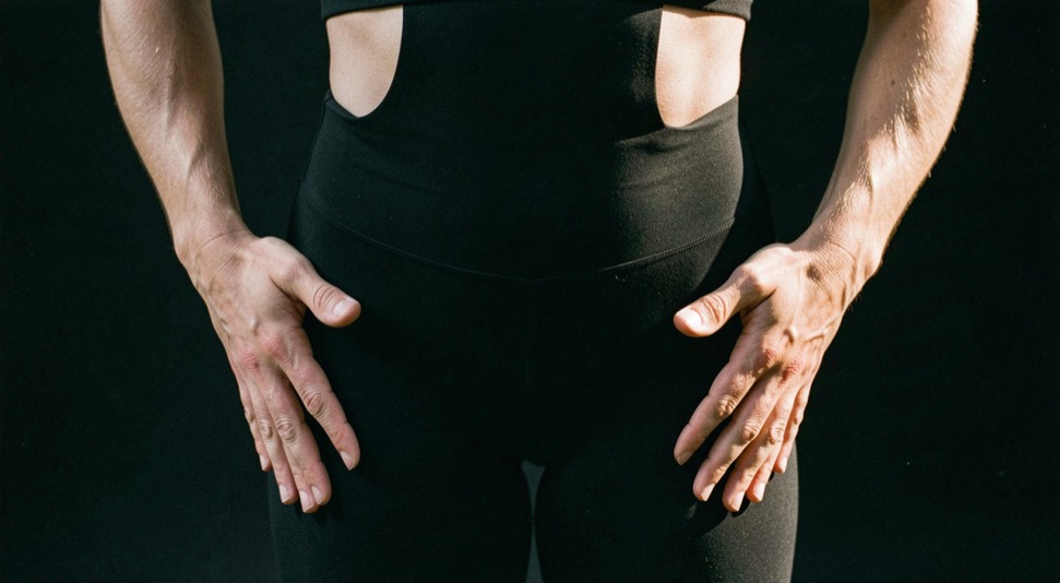 Mains d’une danseuse posées de chaque côté de ses hanches pour contrôler l’immobilité du bassin pendant un exercice, tenue de travail sombre, sol noir, lumière rasante, cadrage vertical serré