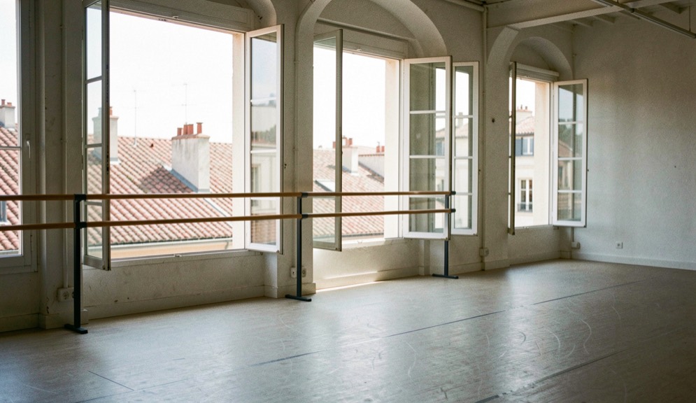 Studio de danse marseillais aux grandes fenêtres ouvertes sur des toits de tuiles, barre le long du mur, sol vide, lumière de midi, cadrage vertical